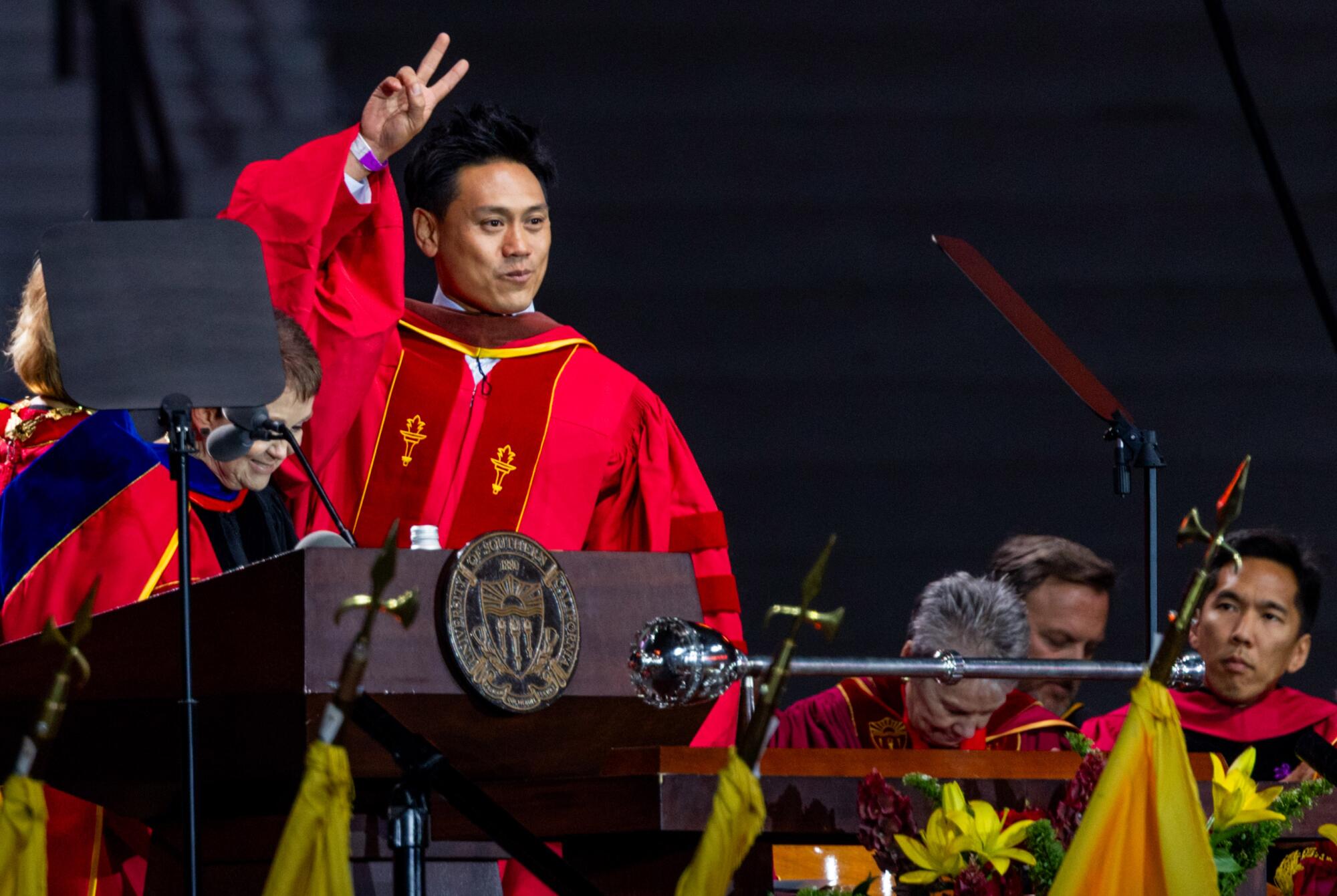 Filmmaker Jon M. Chu in a red academic gown speaks at the USC commencement.