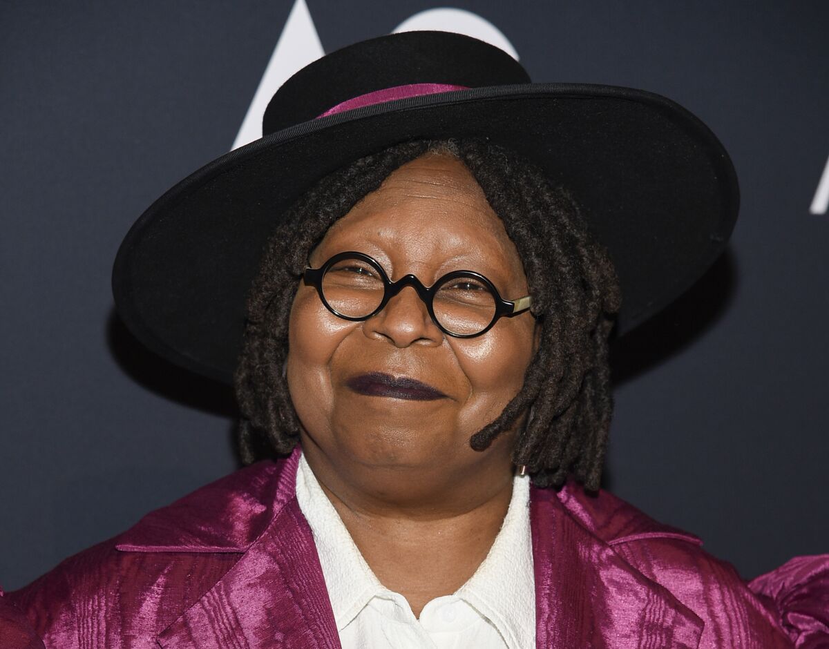 Whoopi Goldberg smiles for the camera in a black hat and magenta outfit