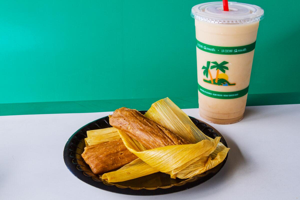 A photo of a plate of two pumpkin spice tamales unwrapped from their husks. Behind is a cup of pumpkin spice horchata.