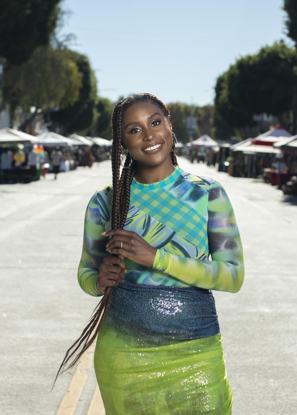 Issa Rael stands in the middle of a street, holding her long braided hair and wearing a bright green patterned outfit