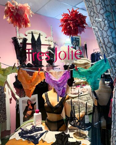 Colorful panties hang on a string with bras on a display table behind a window that says "Tres Jolie."