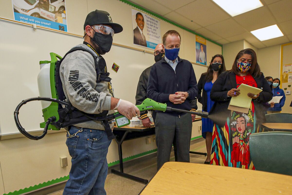 School district officials in a classroom watch a man in protective equipment spray aerosol from a device.