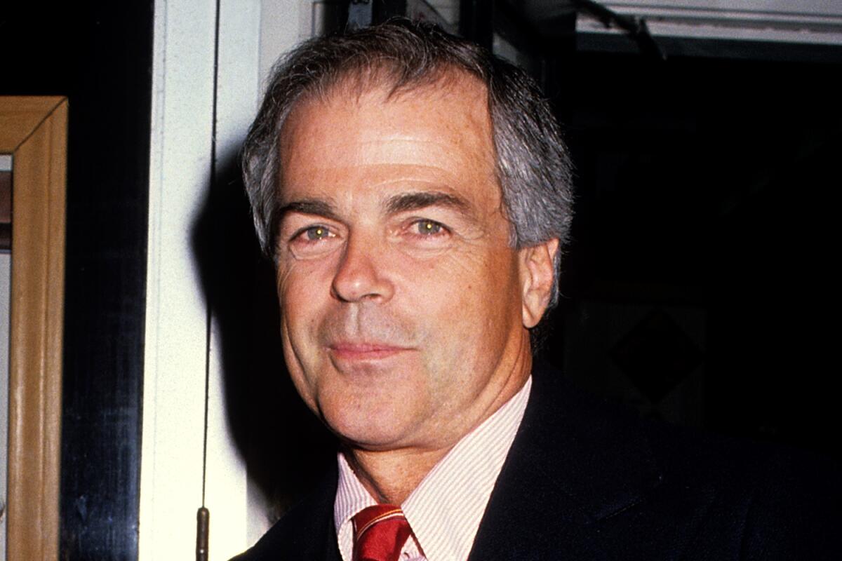 Robert Logan smirking wearing a red tie and white shirt. 