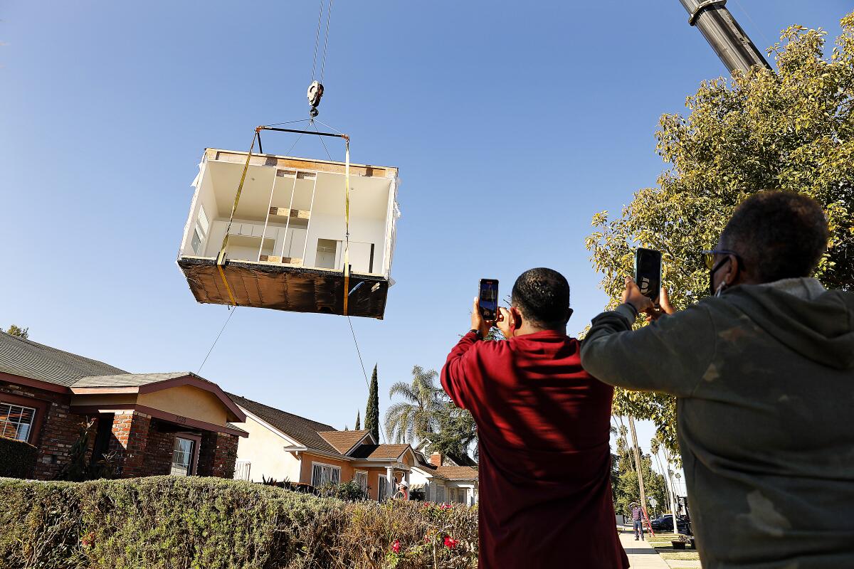 Two people hold up phones to get video of part of a small home being lowered into a backyard with a crane.