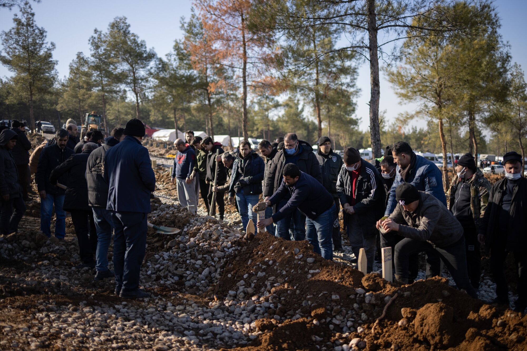 A small crowd stands by as people place wood markers in the freshly turned dirt of a mass grave