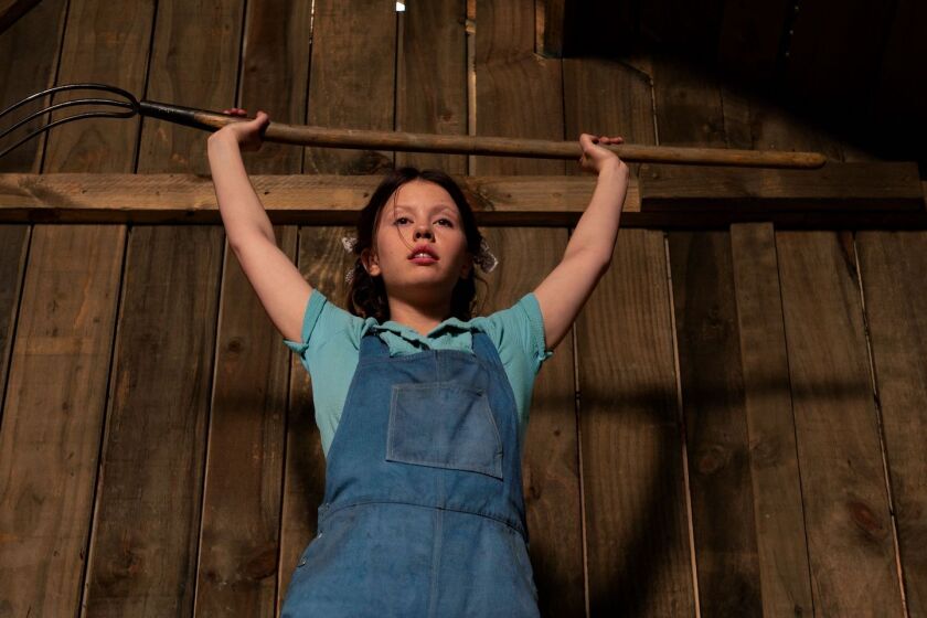 A young woman in overalls raises a pitchfork over head in the movie "Pearl."