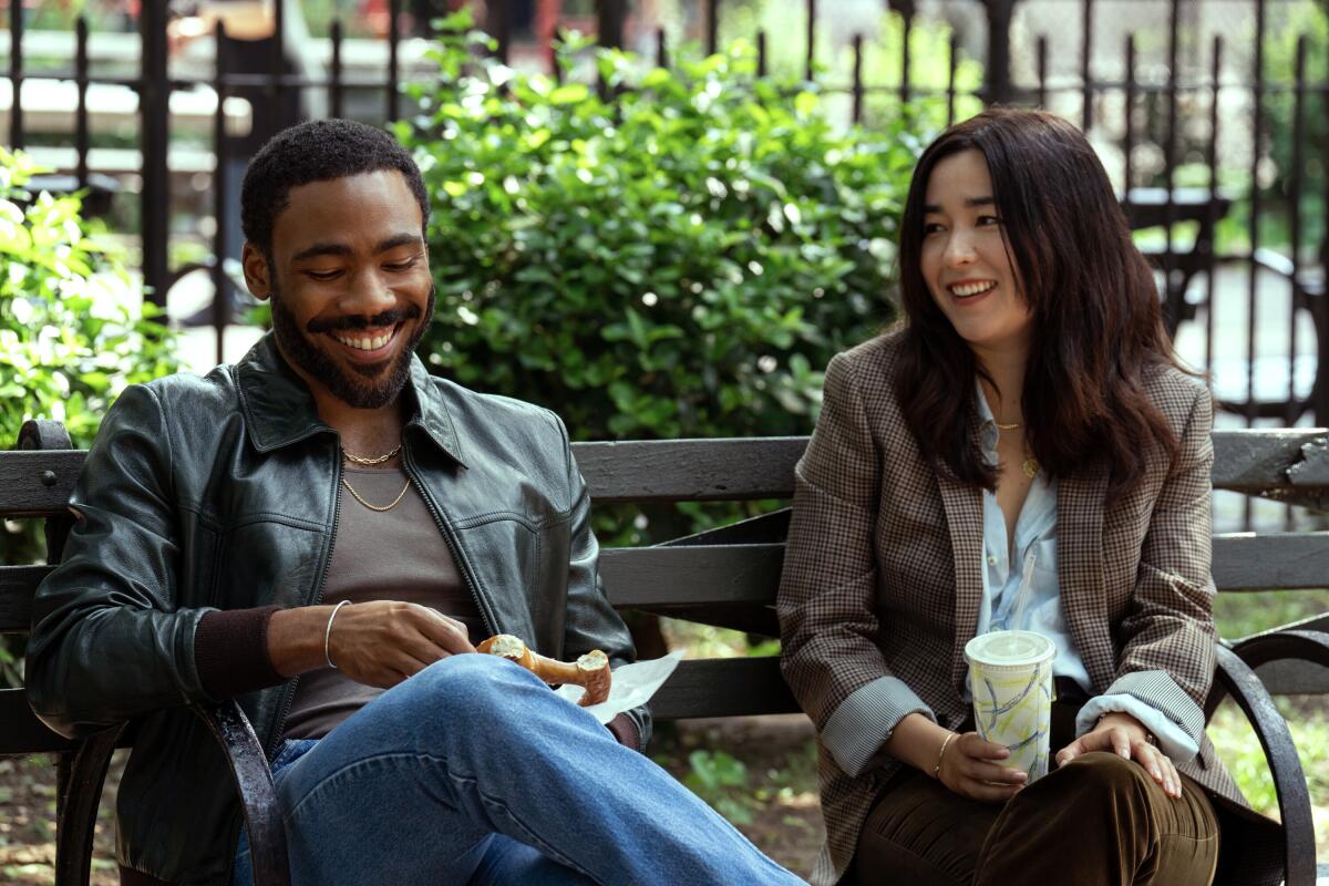 A smiling woman looks at a smiling man who looks at his hands. Both are seated on a park bench.