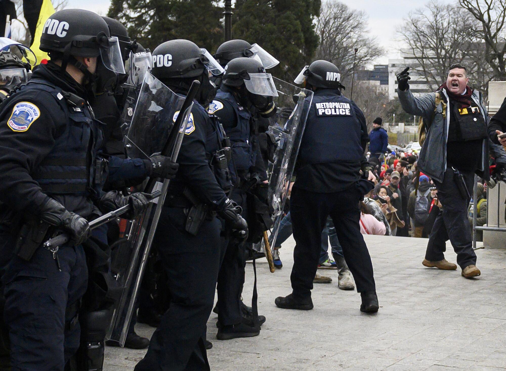 A line of police in riot gear next to a man walking by gesturing with his middle finger toward them