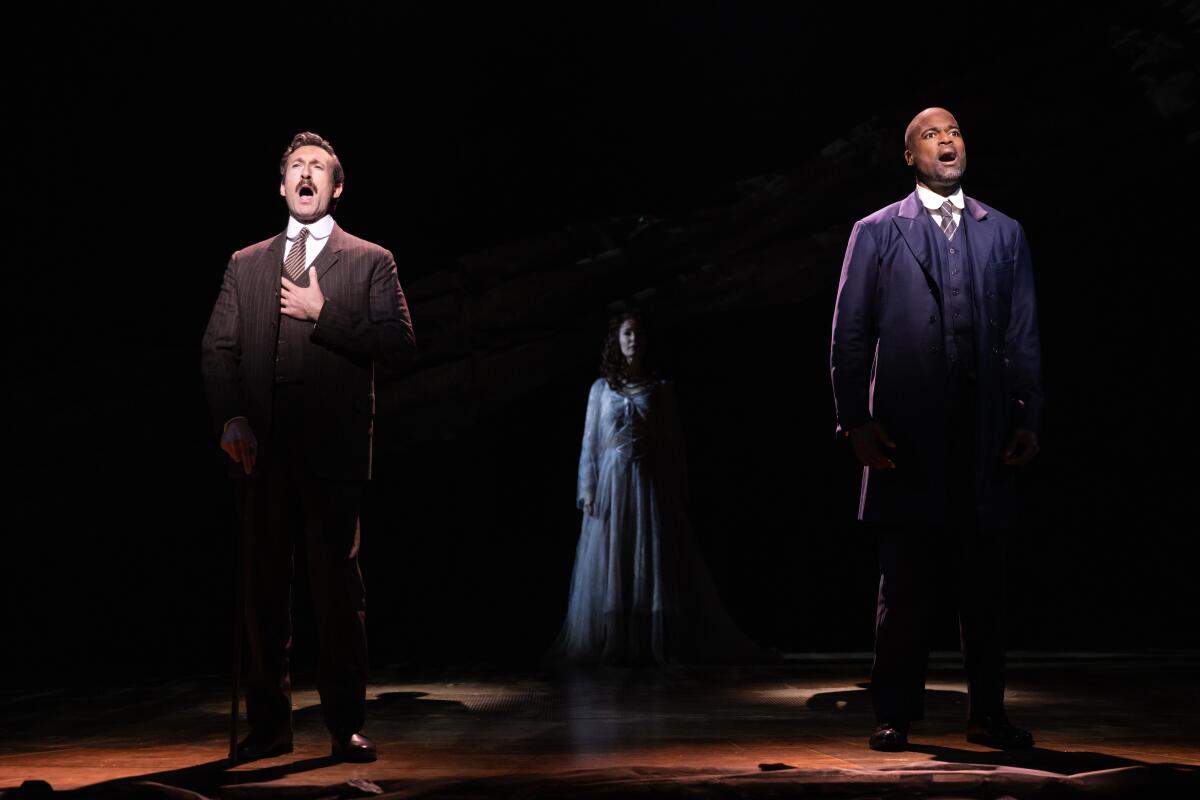 Two actors, spotlighted in the foreground, sing about a figure in half-light behind them.
