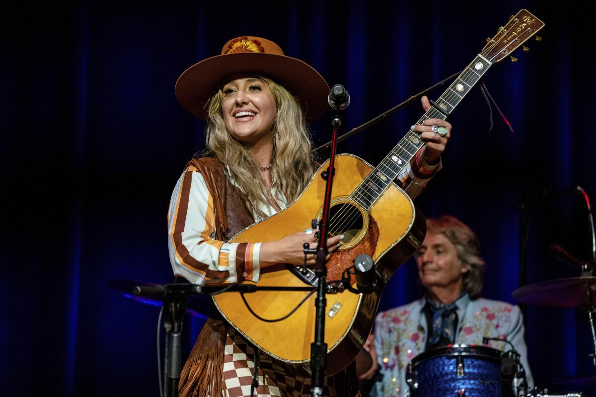 A smiling Lainey Wilson, wearing a large hat, sings and plays guitar onstage