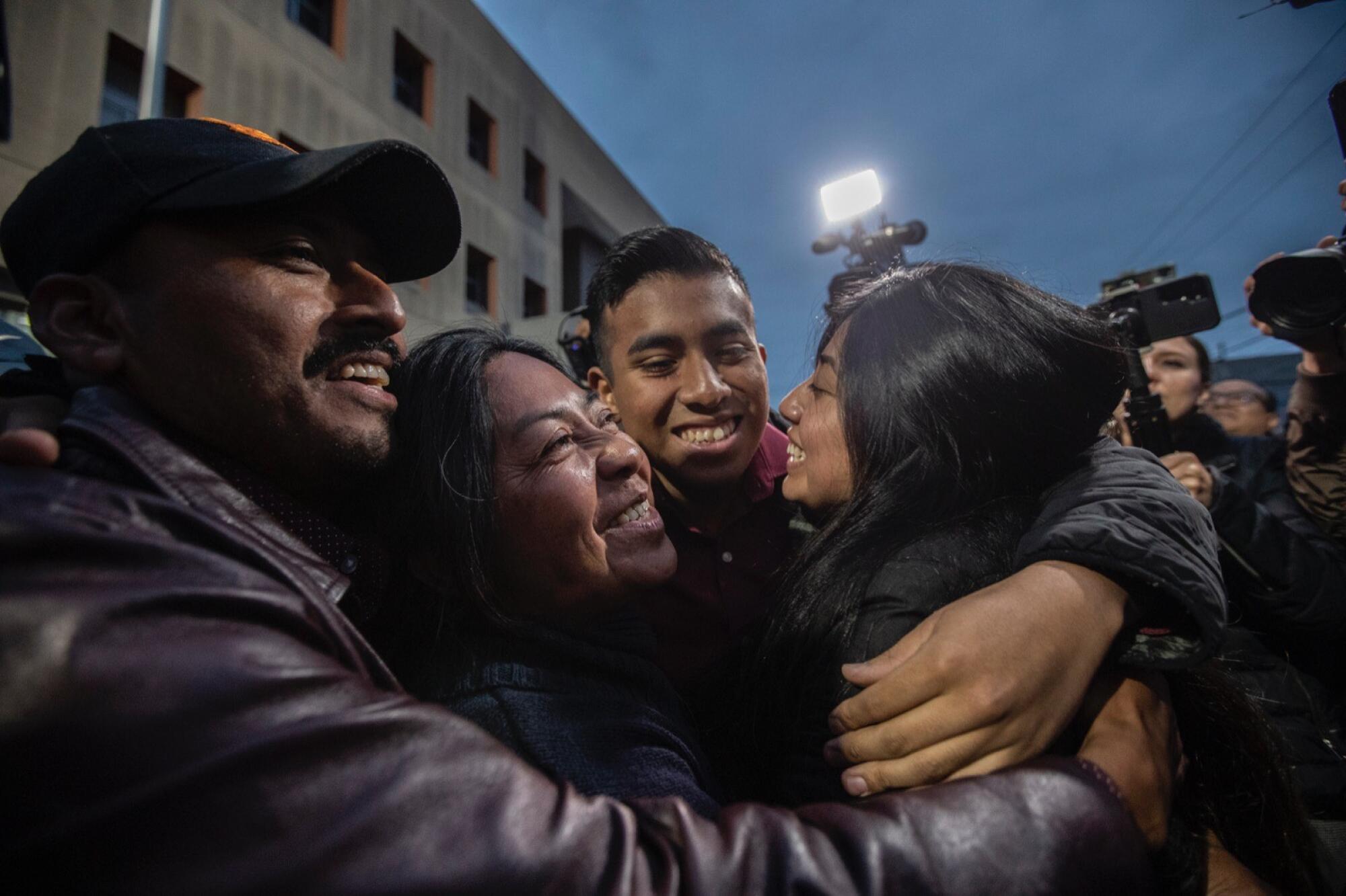 A woman with dark hair is embraced by two men and a woman, all smiling