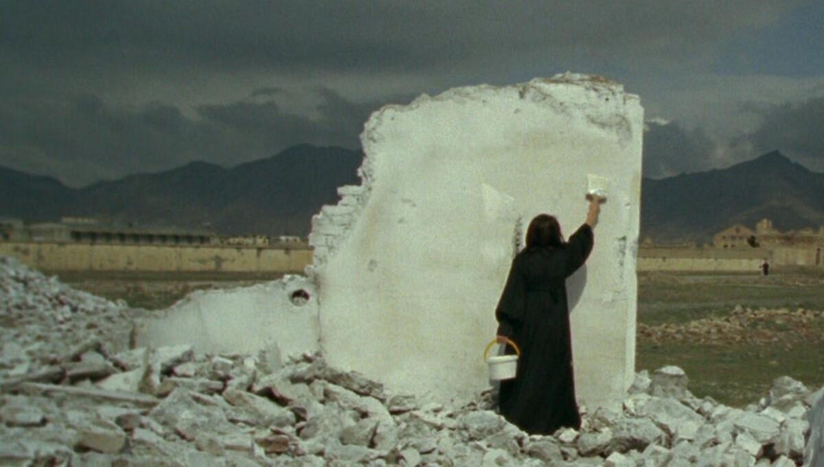 A video still shows a woman in a black tunic standing on a pile of rubble and painting a fragment of wall white.