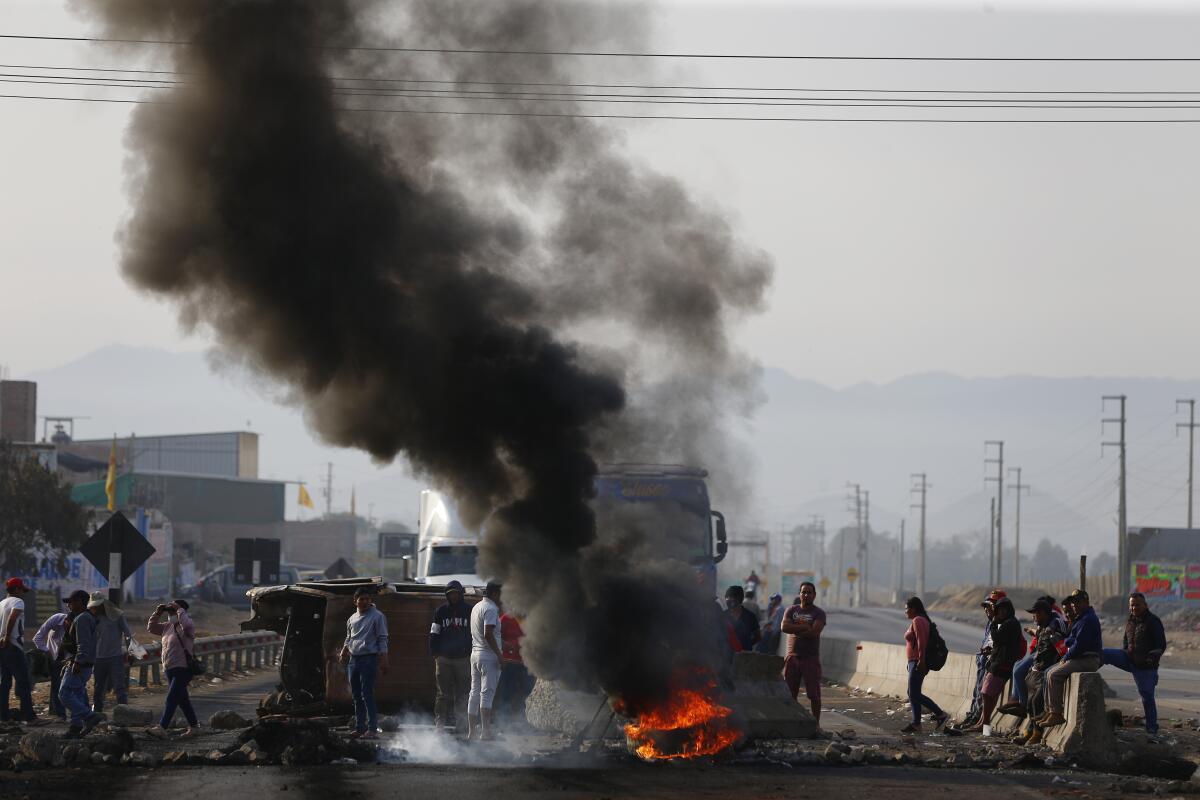 Dark smoke rising from a fire next to a highway as a small crowd looks on.