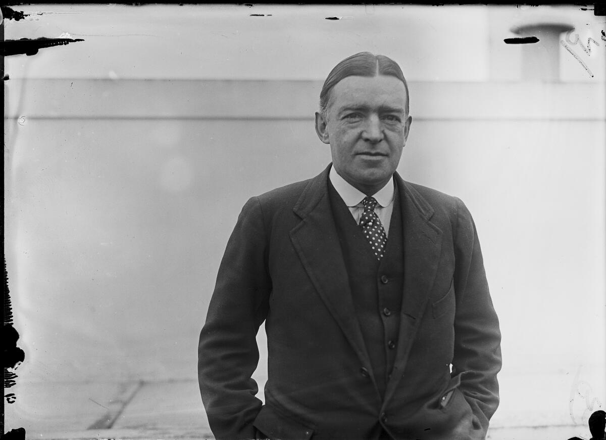 A black-and-white photo of a man in suit and tie standing on a ship