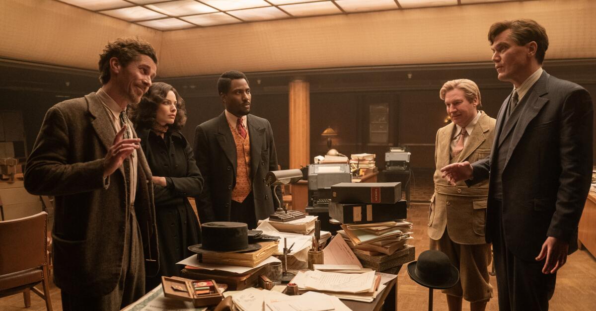 Four men and a woman in period clothing stand around a table with piles of papers and books.