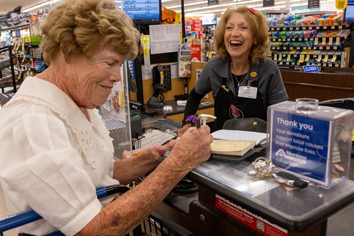A grocery store cashier and customer smile as the older woman writes a check for her groceries.