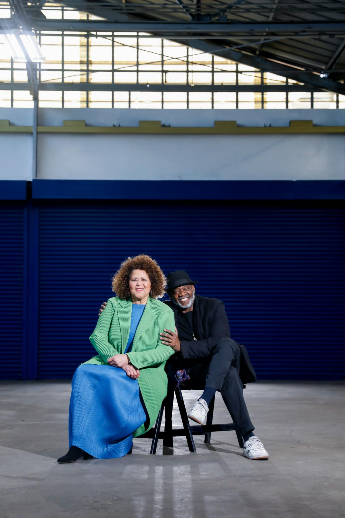 Anna Deavere Smith, in blue dress and green coat, shares a bench with Gregg Daniel, in black, in a bare industrial space