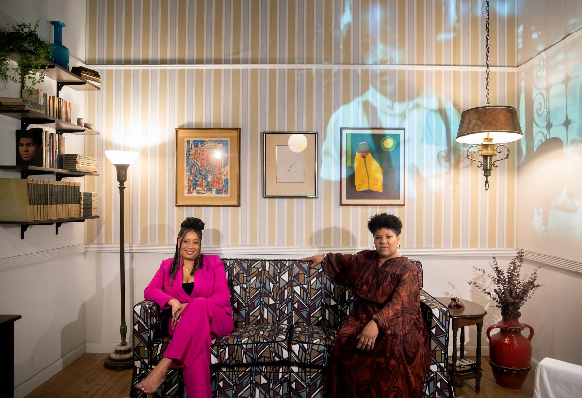 Two women sit in a wallpapered living room. 