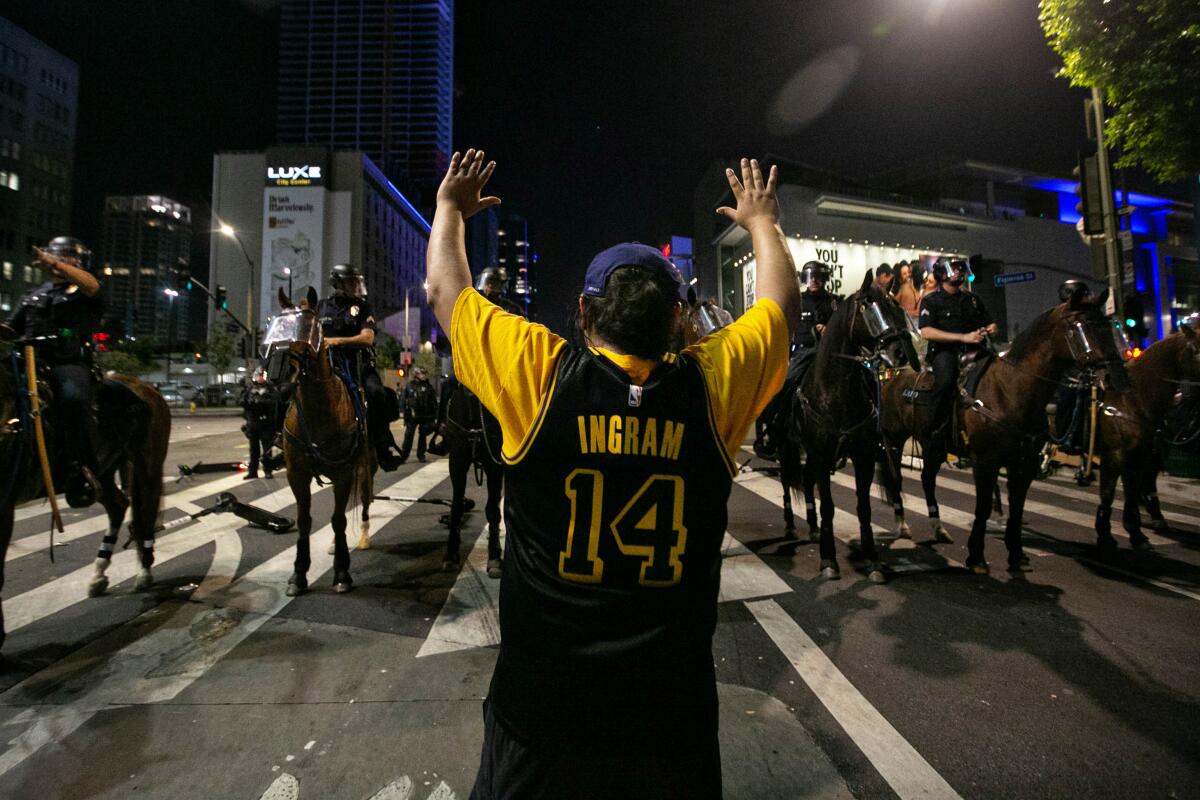 A man in a black Ingram 14 Lakers jersey raises his arms in front of a line of police officers on horseback