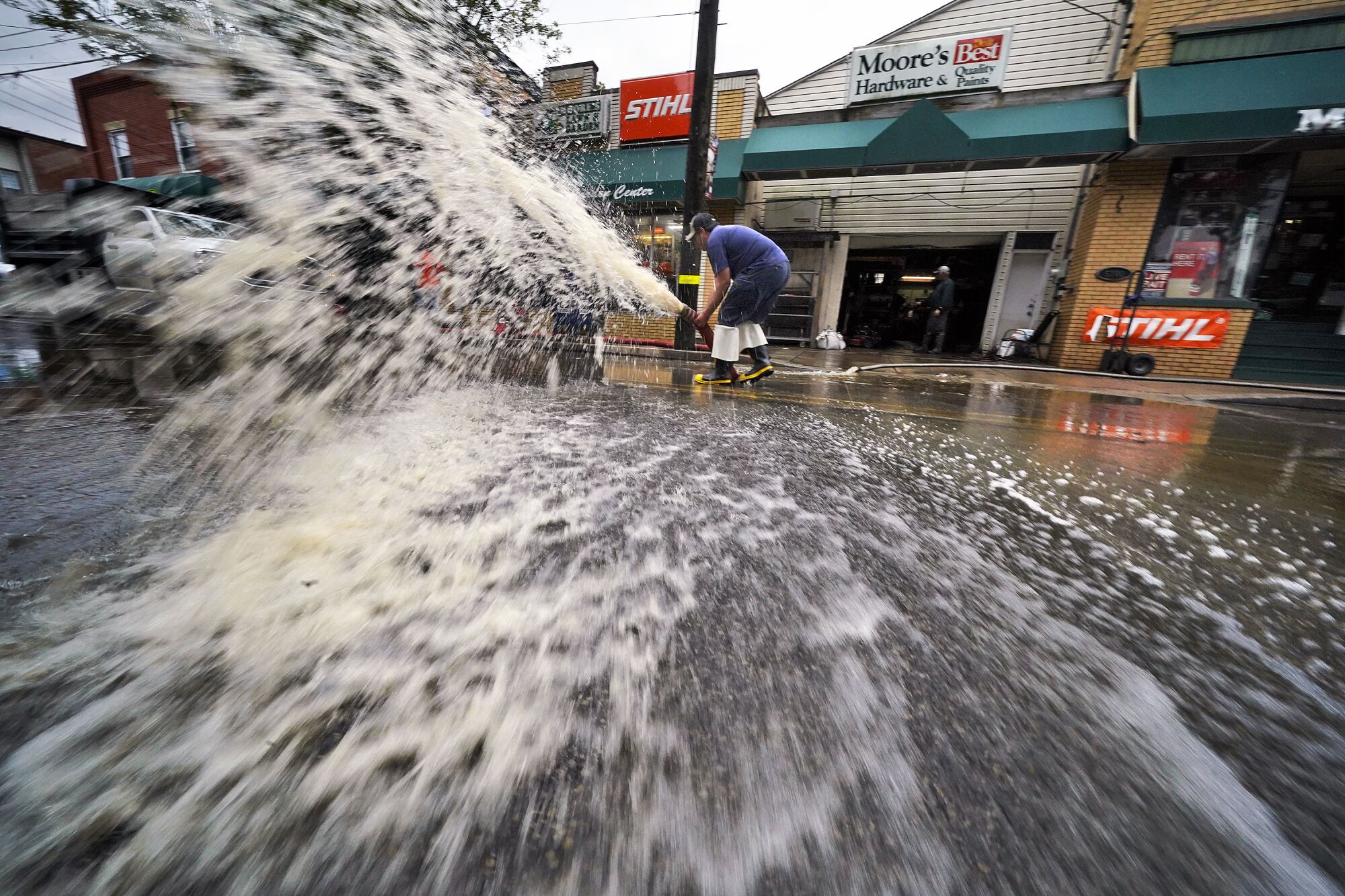 Water sprays dramatically from a pump on a street outside a hardware store.
