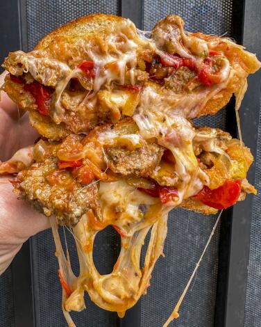 A hand holds up two halves of a sausage and peppers sandwich dripping with melted cheese