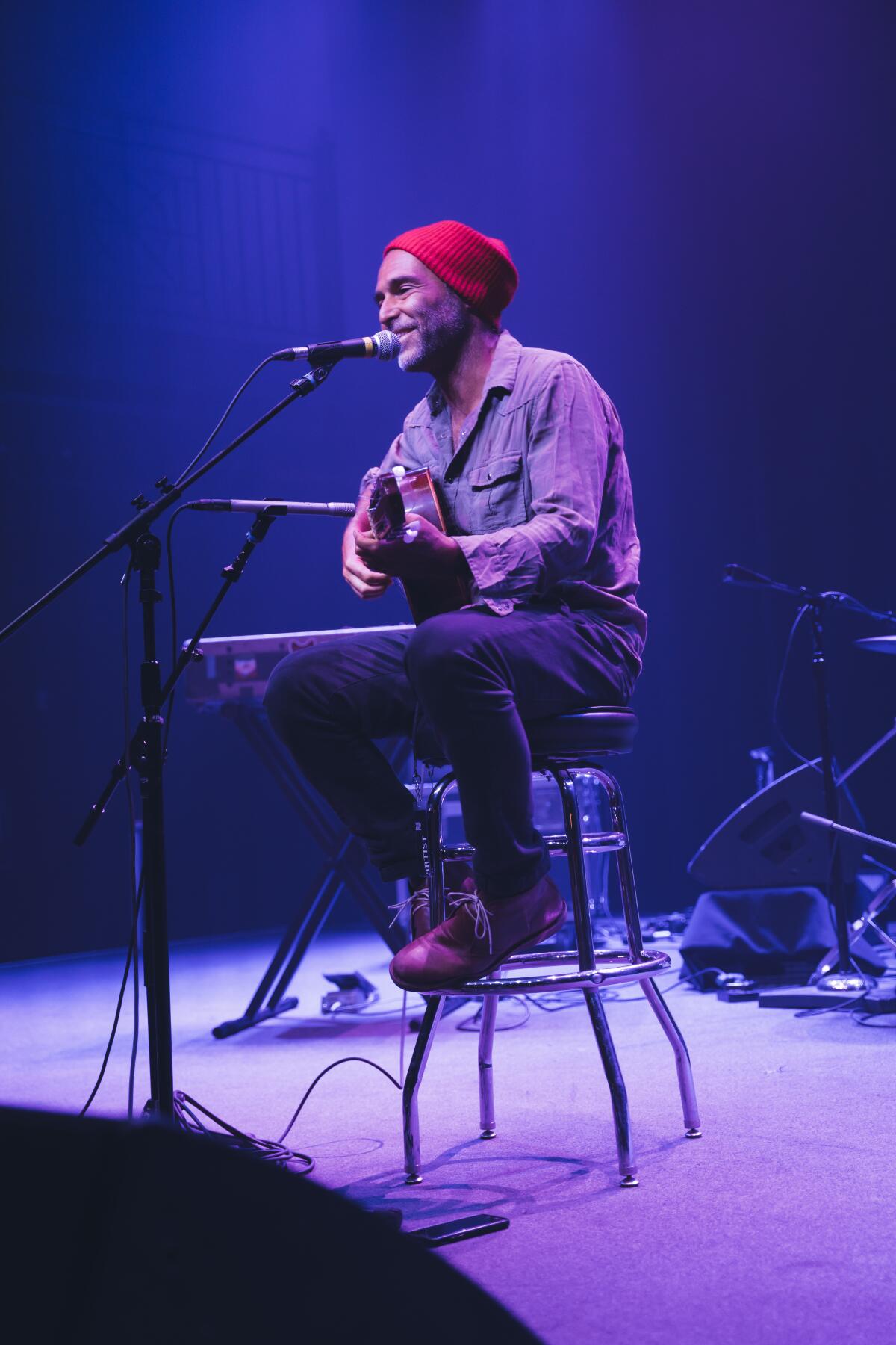A man in a red wool cap seated on a stool sings and plays acoustic guitar.