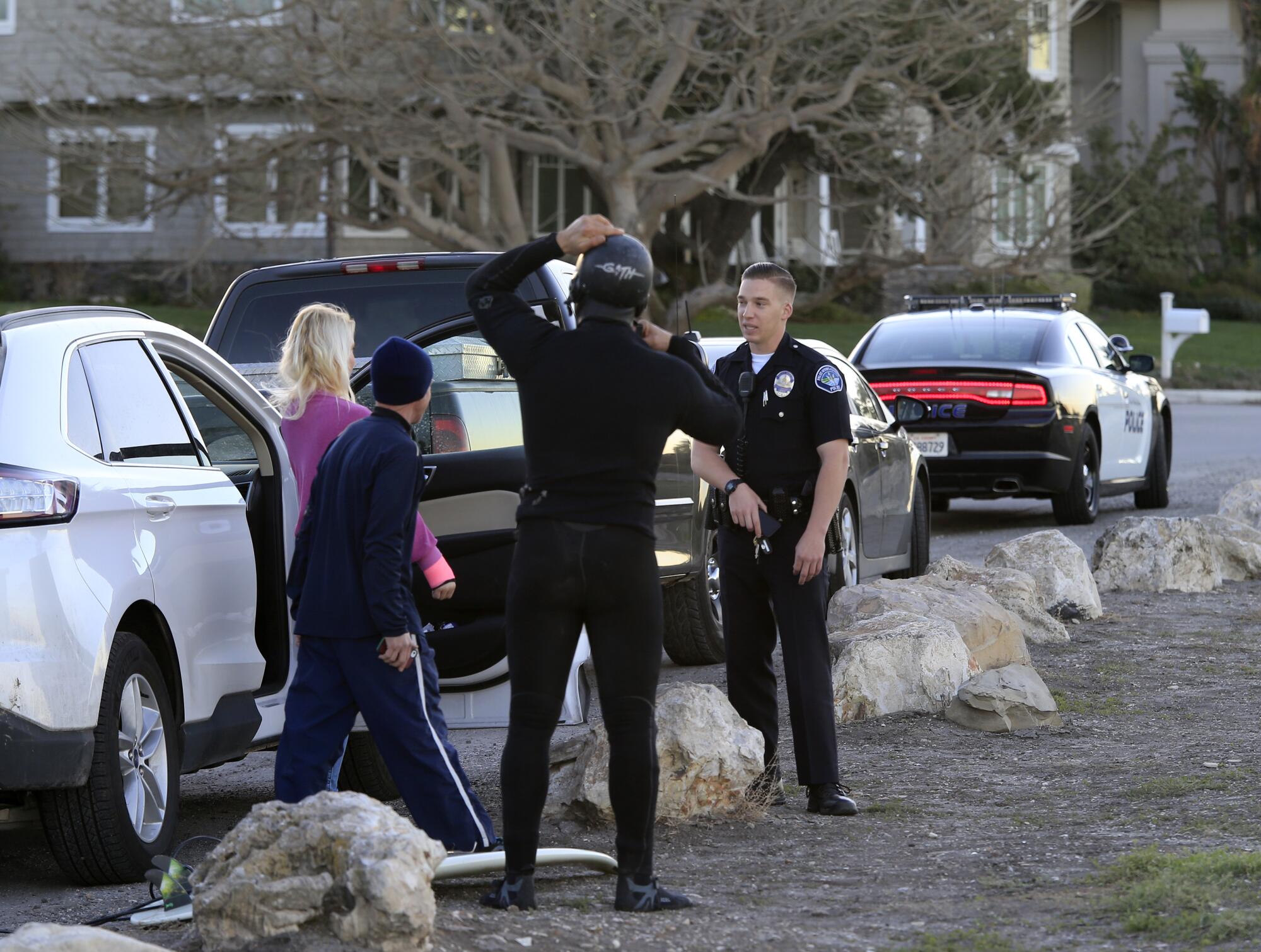 Three people face a police officer next to parked cars.