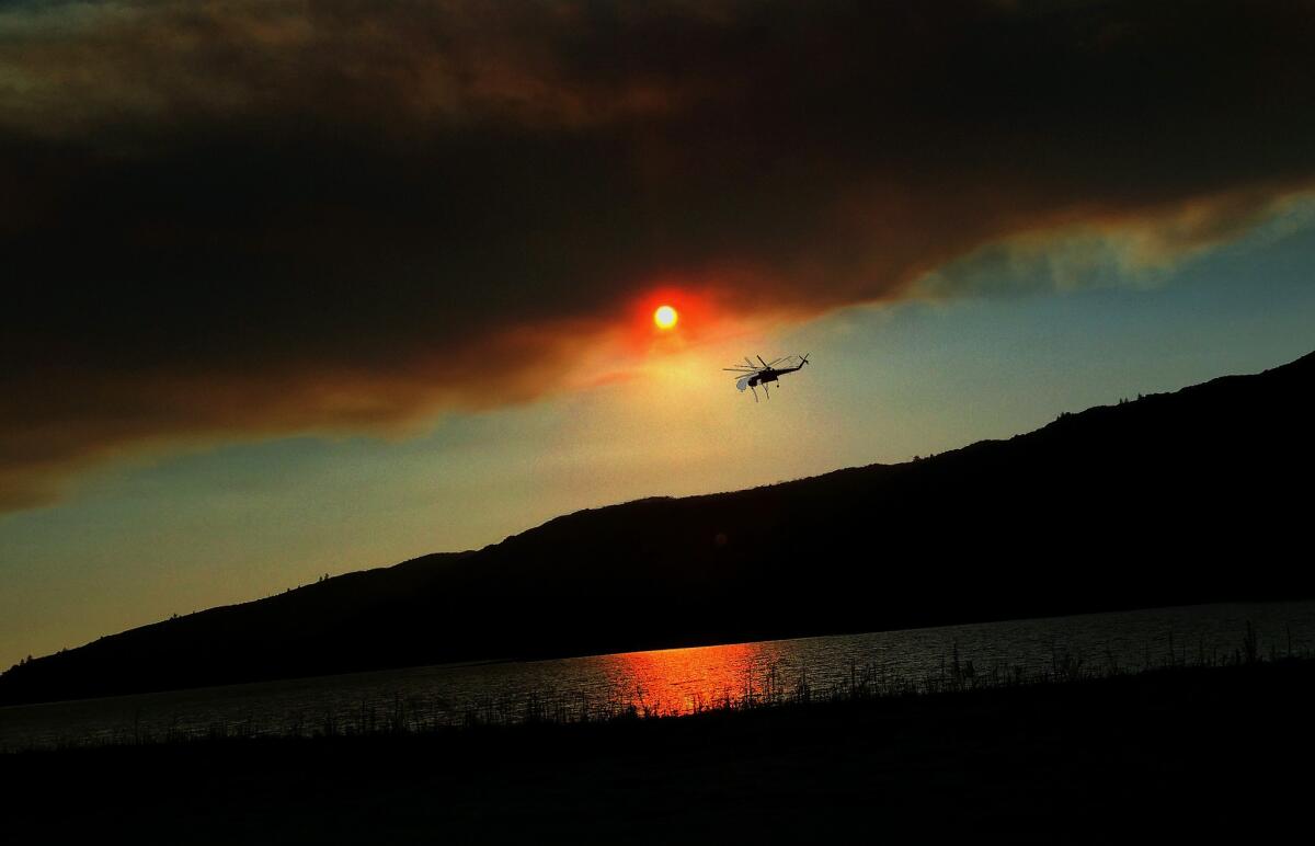A plume of smoke looms over Lake Hemet in front of the setting sun as a water-dropping helicopter heads to the lake for a refill while battling the Mountain fire.