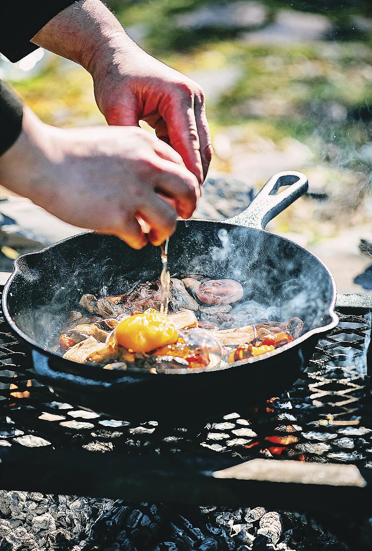A person cooks breakfast in a cast-iron skillet over a campfire.