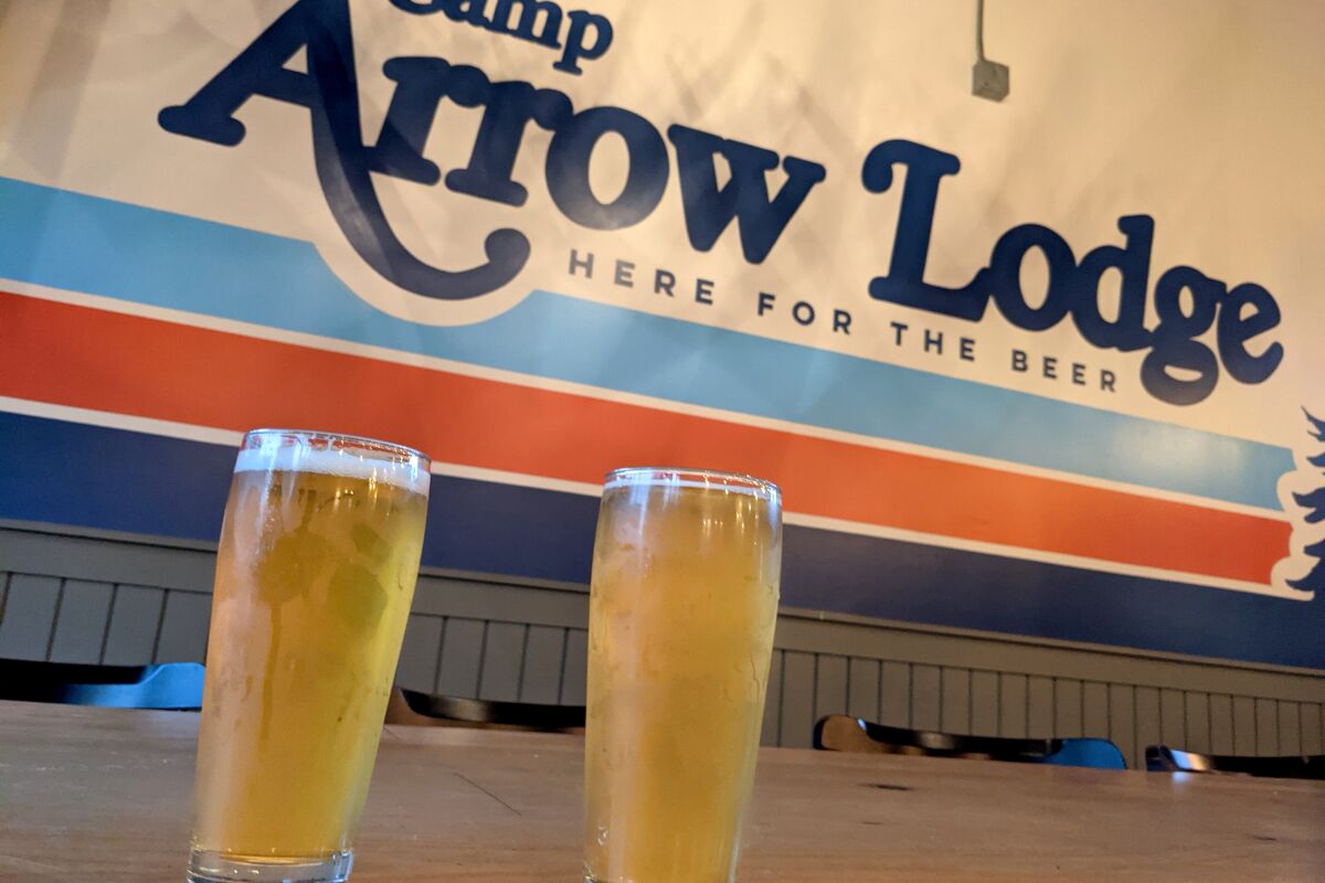 Two beers on a table in front of a sign that says "Camp Arrow Lodge Here for the Beer"