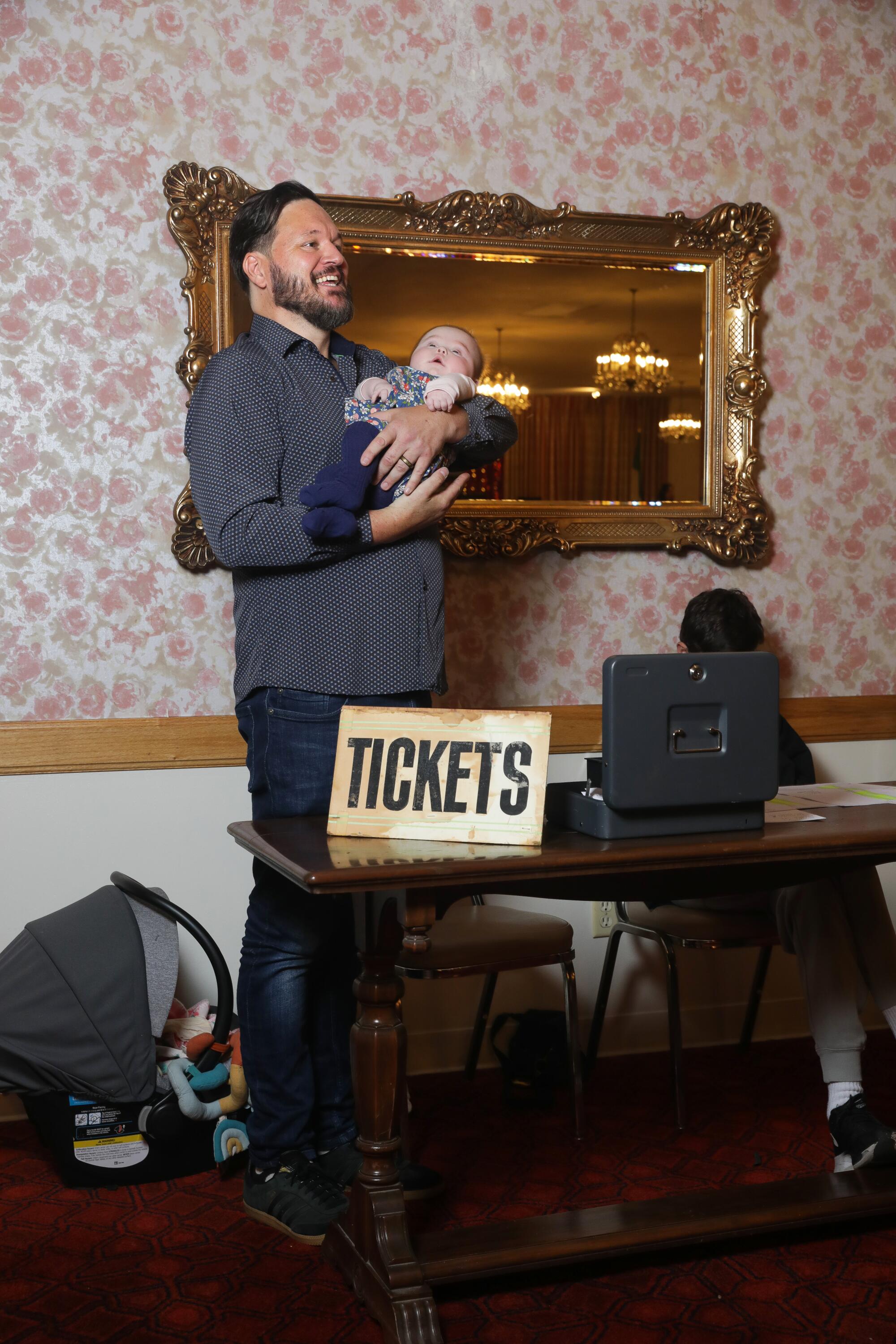 A man holds his baby behind a table with a sign that says "tickets"  at the Garibaldina Society.
