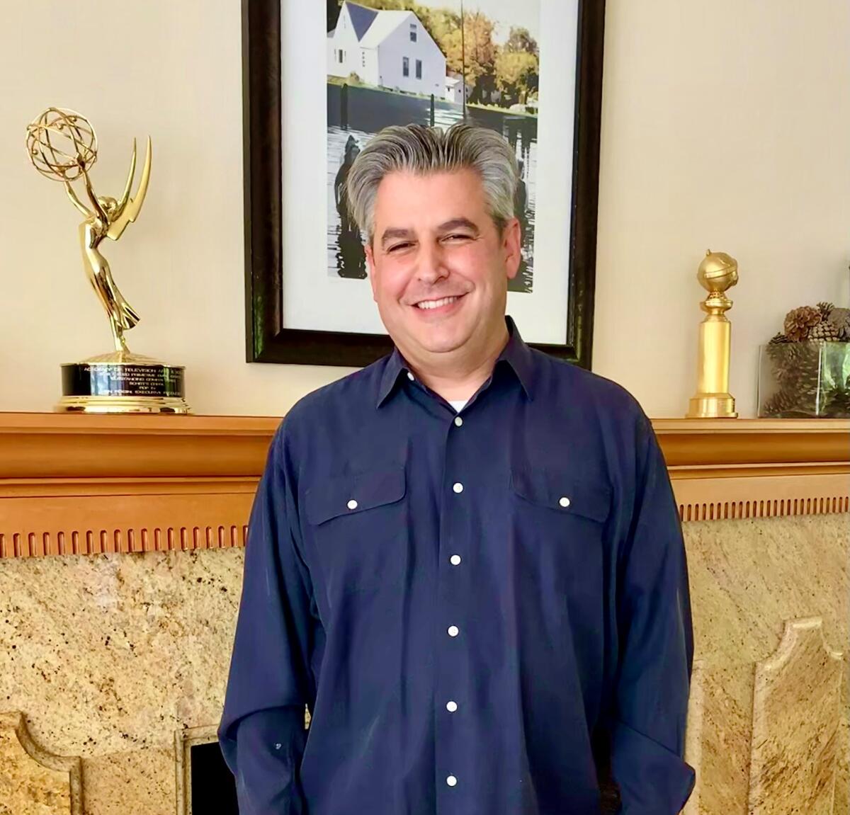 A man with gray hair, smiling in a blue button-up shirt, stands next to a mantel with Emmy and Golden Globe awards on it.