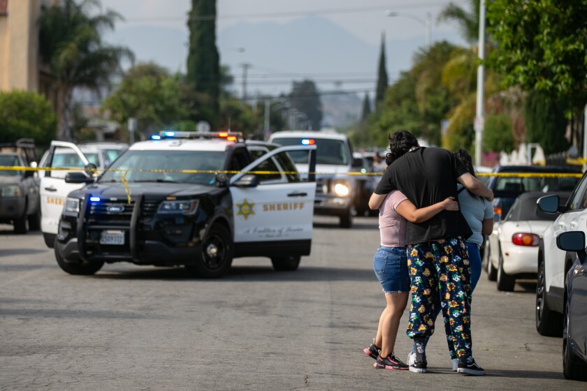 Neighbors embrace in front of an area with police cars and crime scene tape