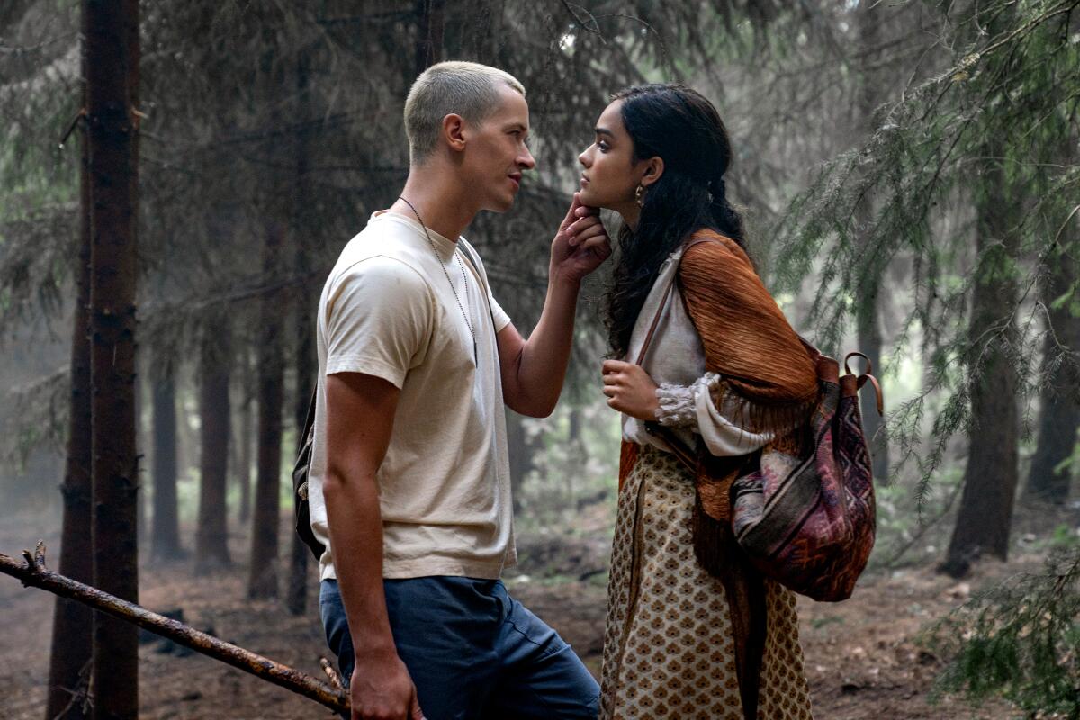 A man in a white shirt and blue jeans stands close to a woman in a sleeveless top in the woods.