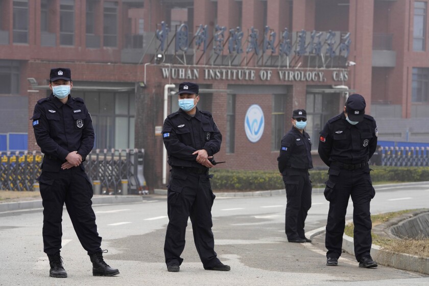 Four men in uniforms and masks stand on a roadway outside a building with a sign "Wuhan Institute of Virology."