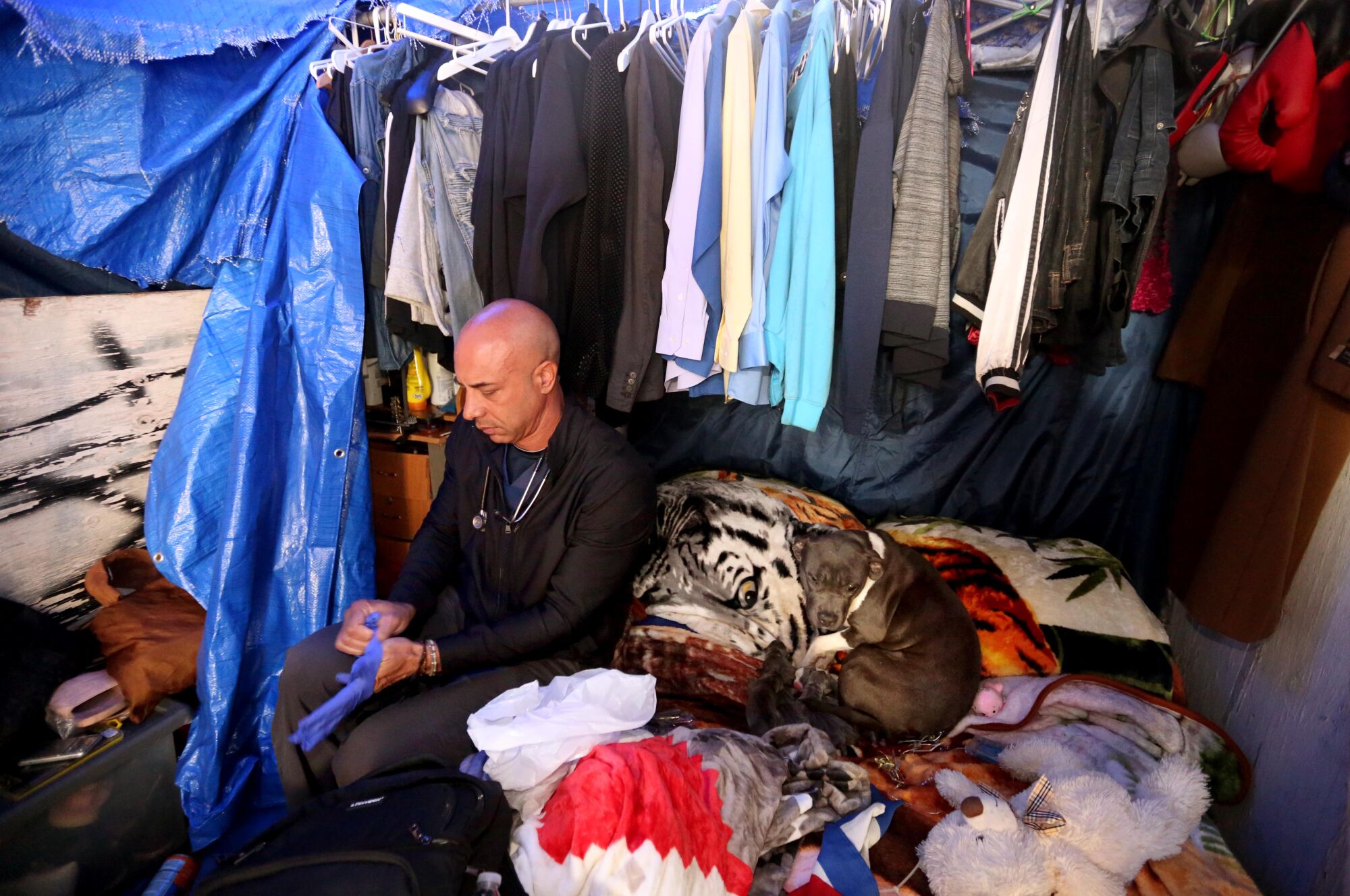 A man sits on a mattress alongside a tarp and clothes hanging from a rod. Beside him on the bed a dog is curled up.