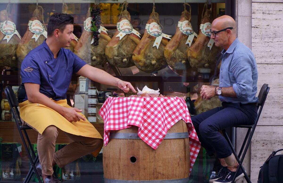 A man in an apron and Stanley Tucci sit on either side of a barrel with a checkered tablecloth.