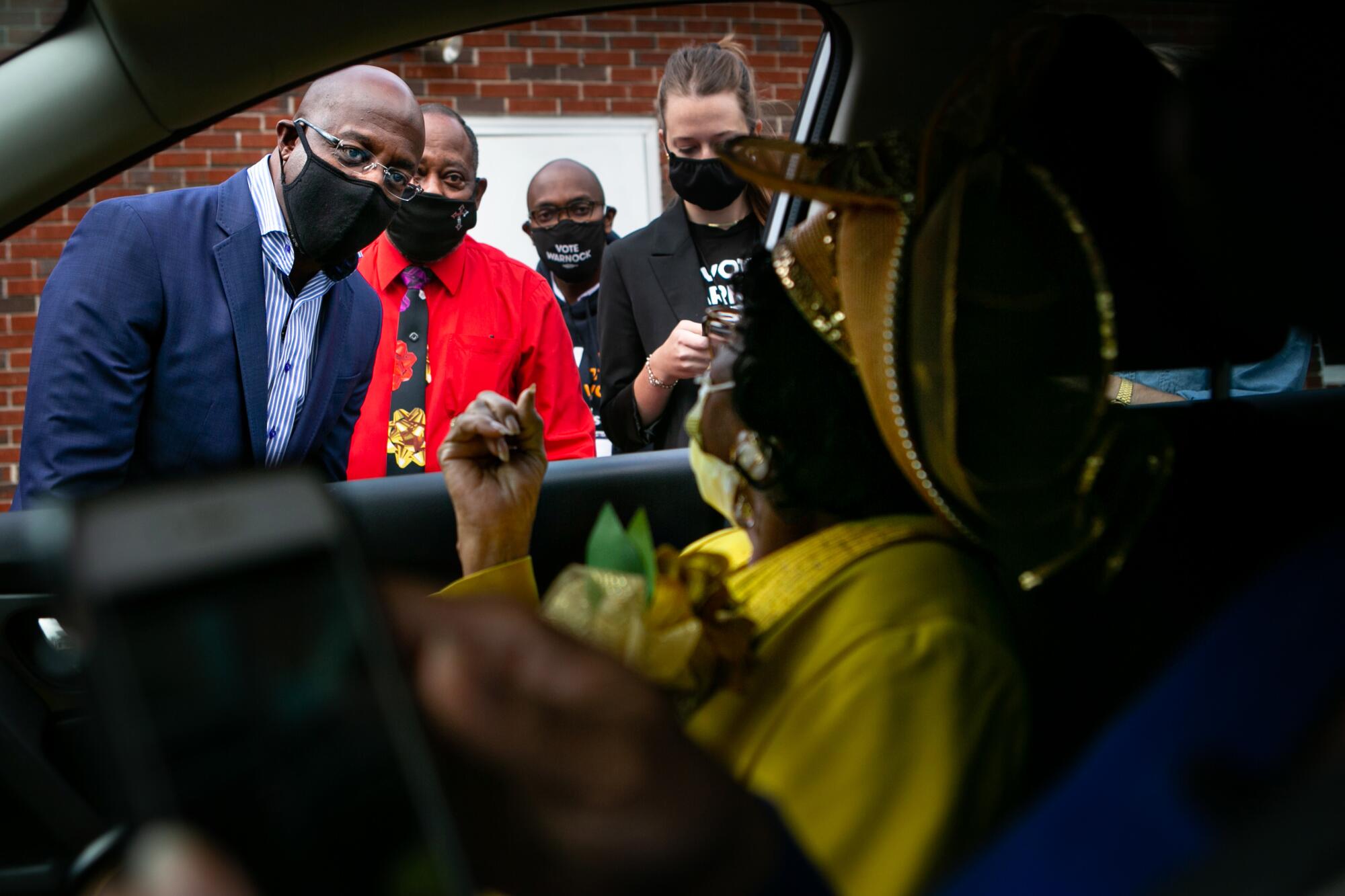 The Rev. Warnock, standing with several others in masks, speaks with a woman inside a car.