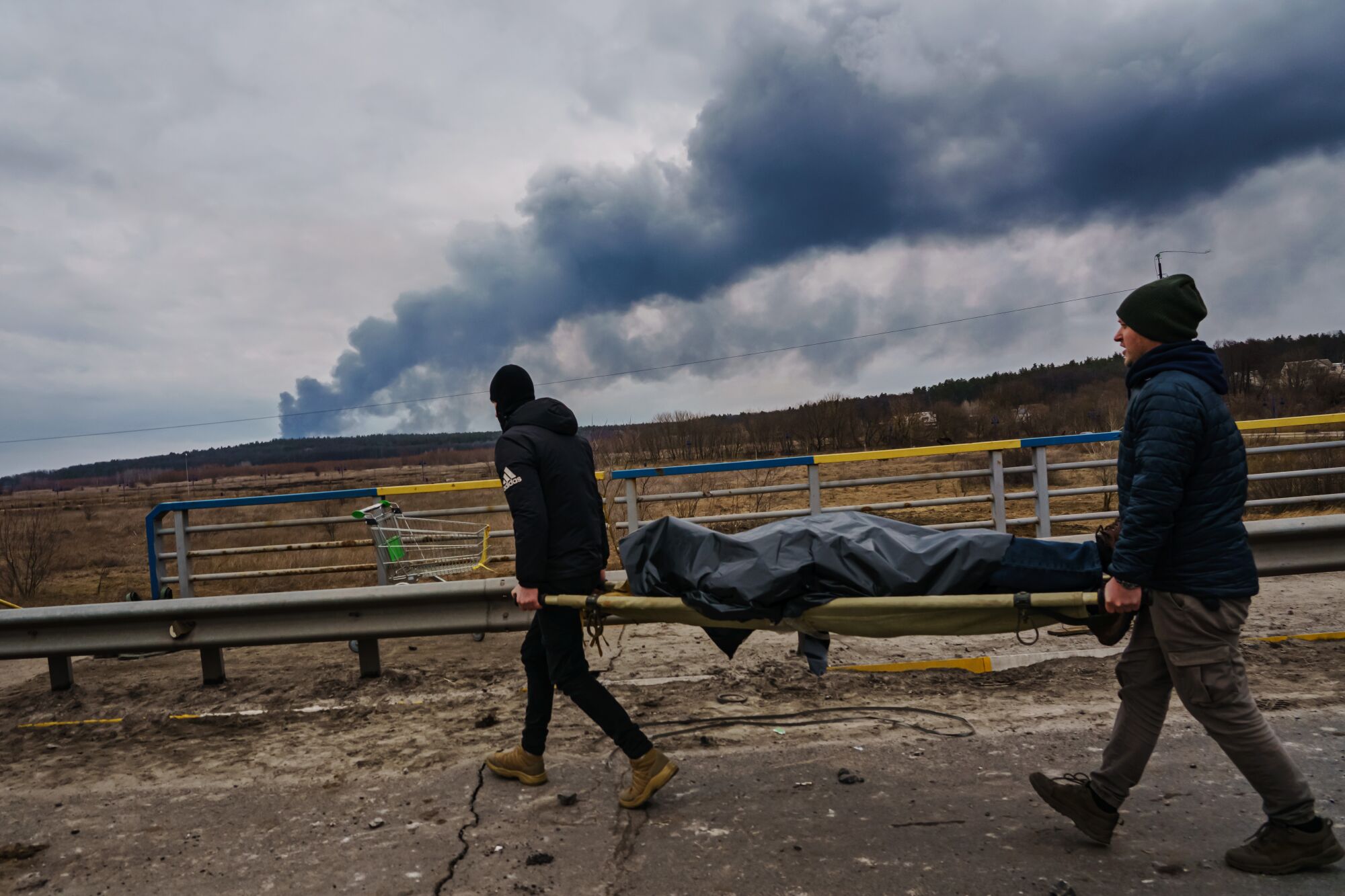 A year of war: Los Angeles Times photographers document the battle in Ukraine 6 Two people carry a stretcher with a body as smoke rises in the distance.