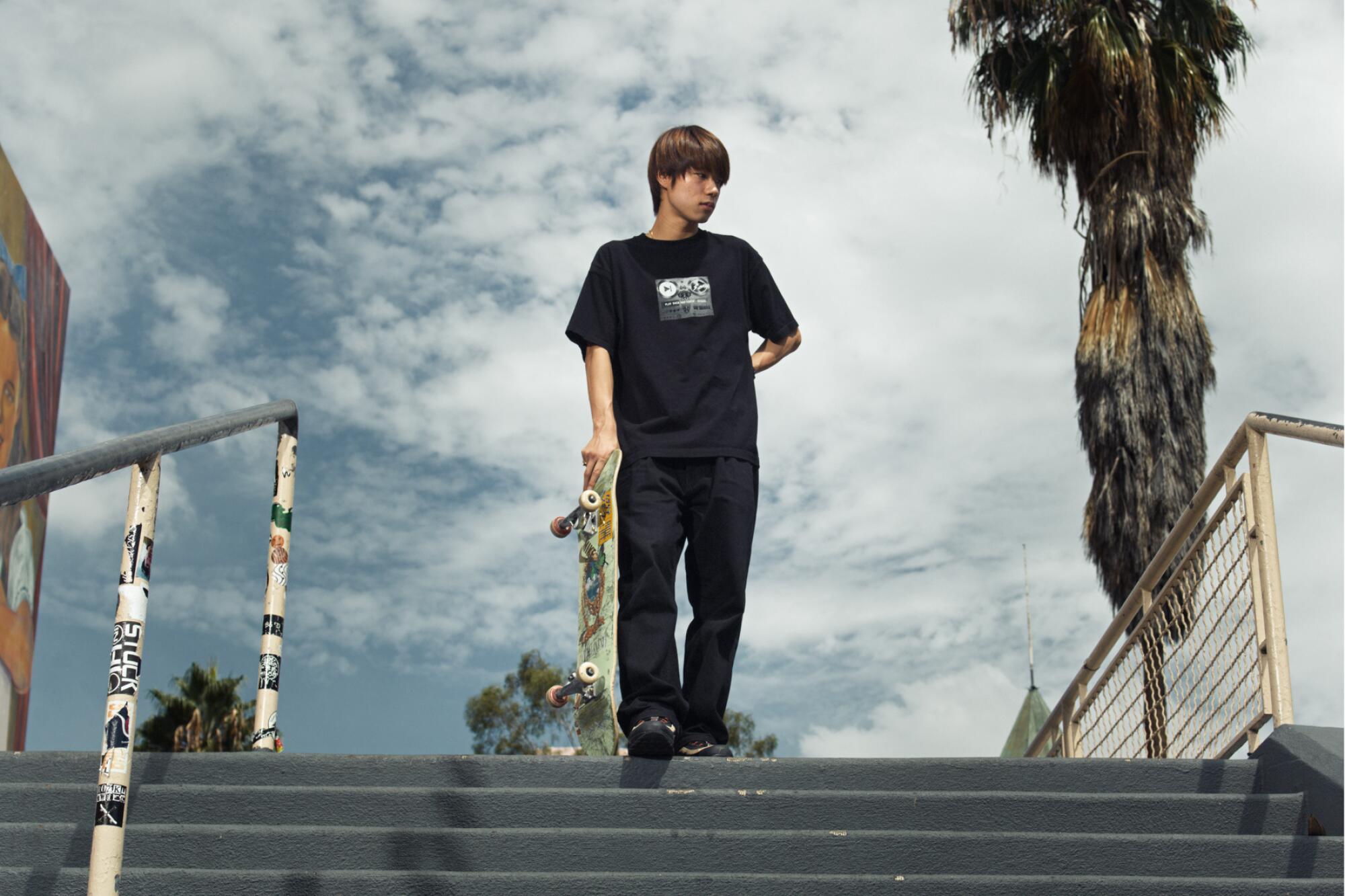 A young man at the top of a staircase holds a skateboard to his side 