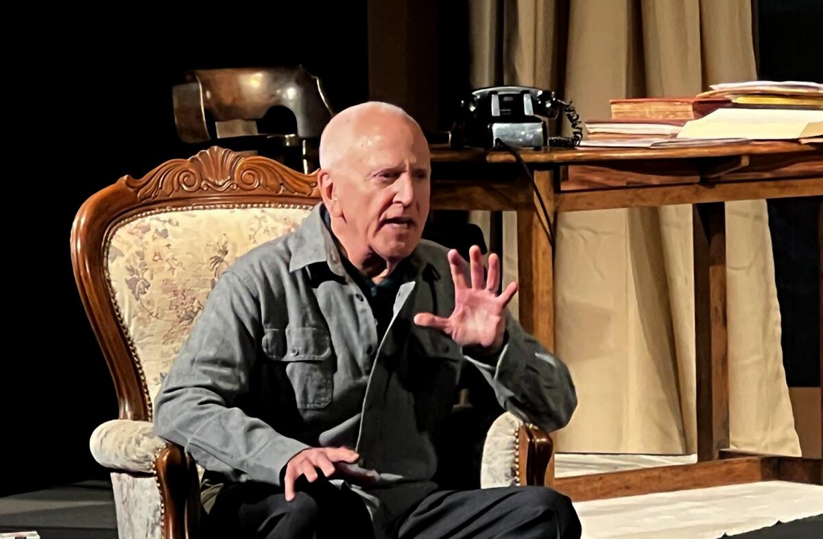 A gray-haired man seated in a chair, talking and gesturing with his hands, onstage.