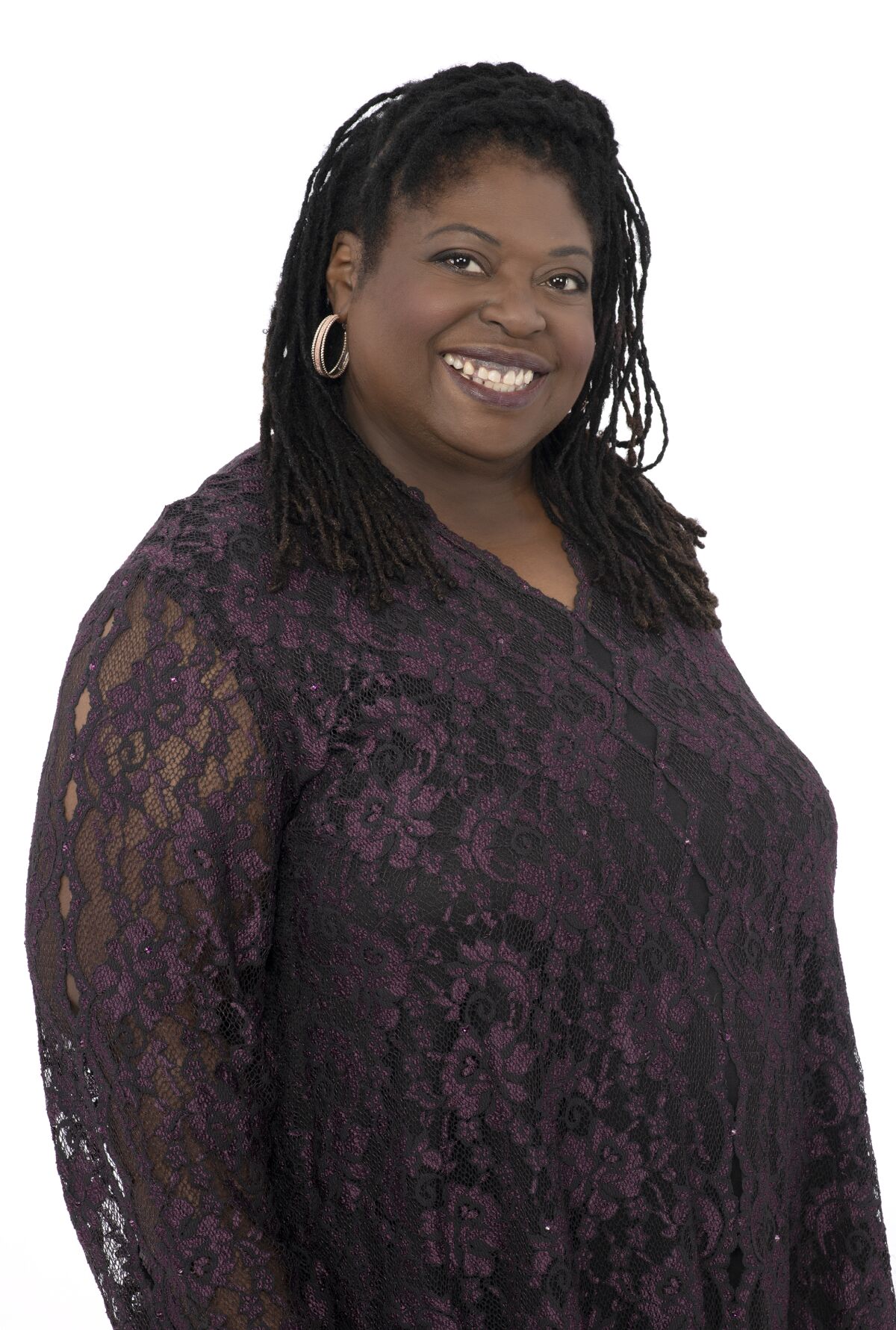 A woman with braids and hoops and wearing a dark-colored lace dress smiles at the camera.