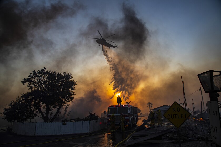 A helicopter drops water on a neighborhood amid dense smoke.