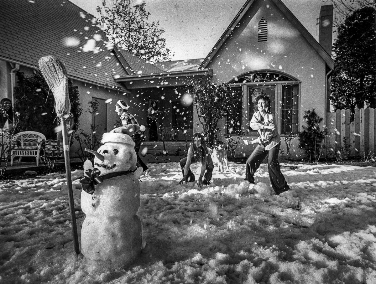 Three children laugh and have a snowball fight behind a snowman in a snow-covered yard in Sherman Oaks.