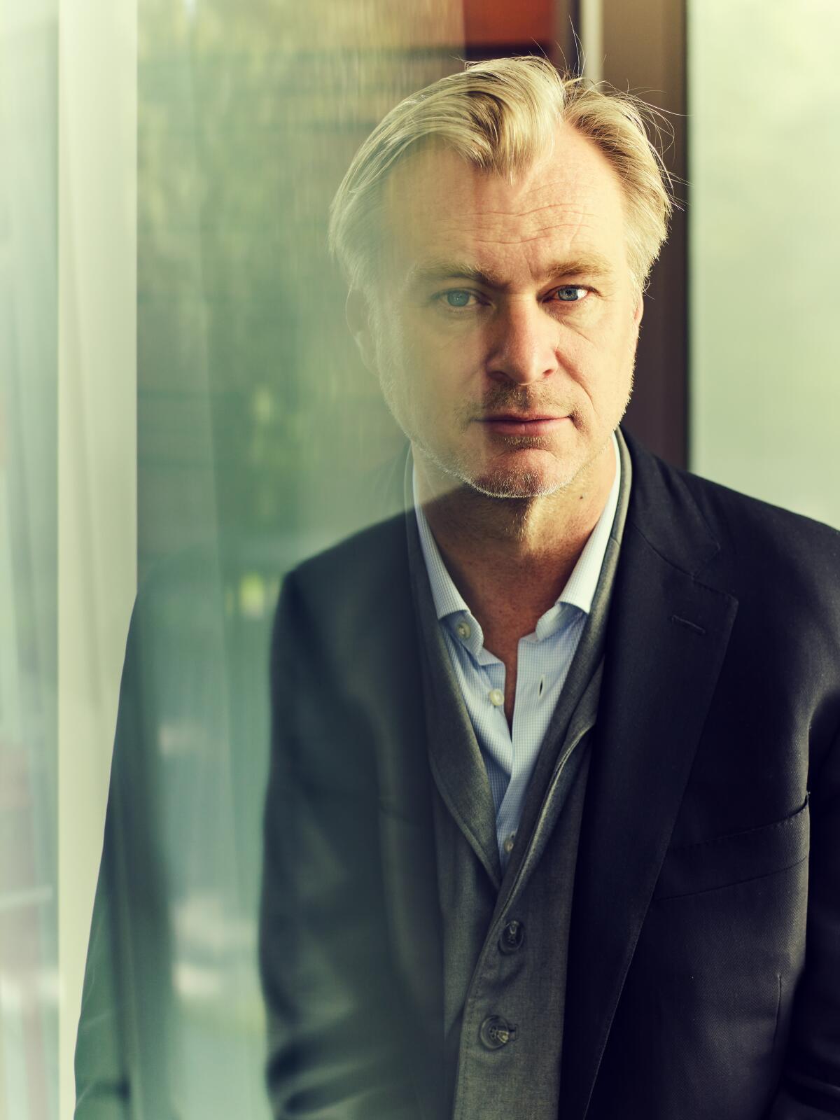 A blond, middle-aged man in a suit with no tie (Christopher Nolan) seen through glass with a translucent reflection 