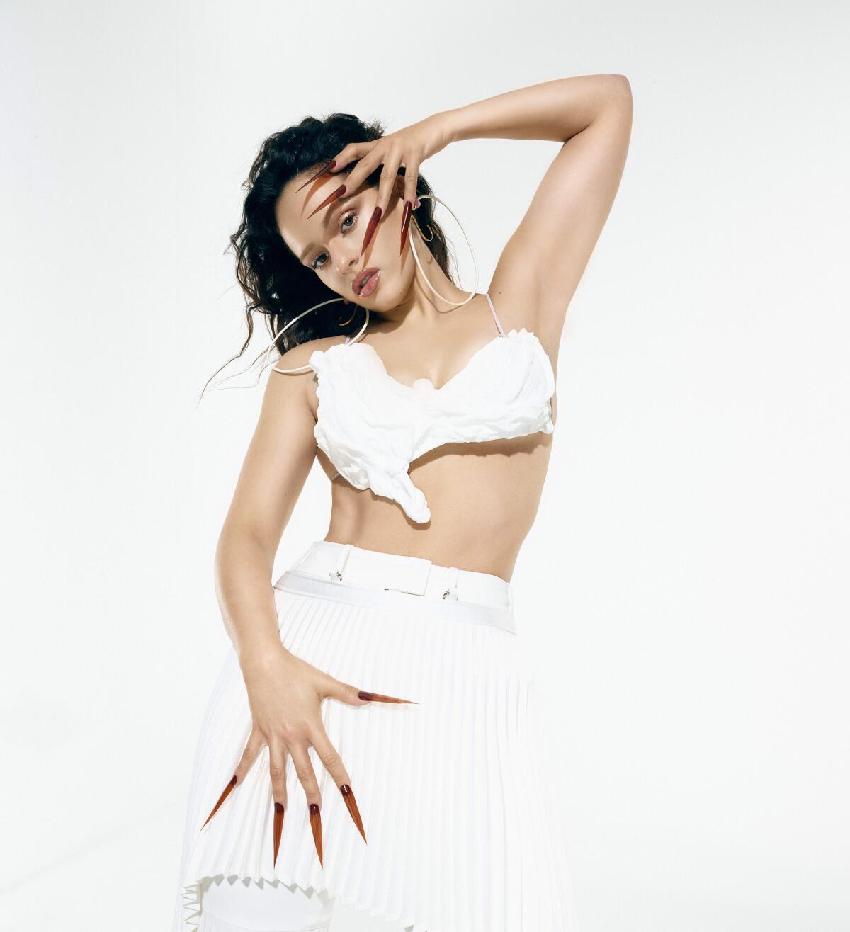 A woman with long sharp fingernails poses in a skimpy white top and skirt 