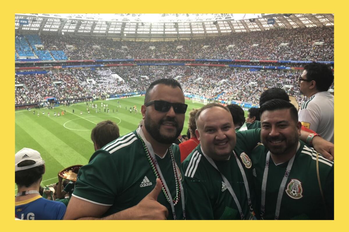 Three men pose for a photo in an upper level of the stands of a soccer stadium with the field in the background