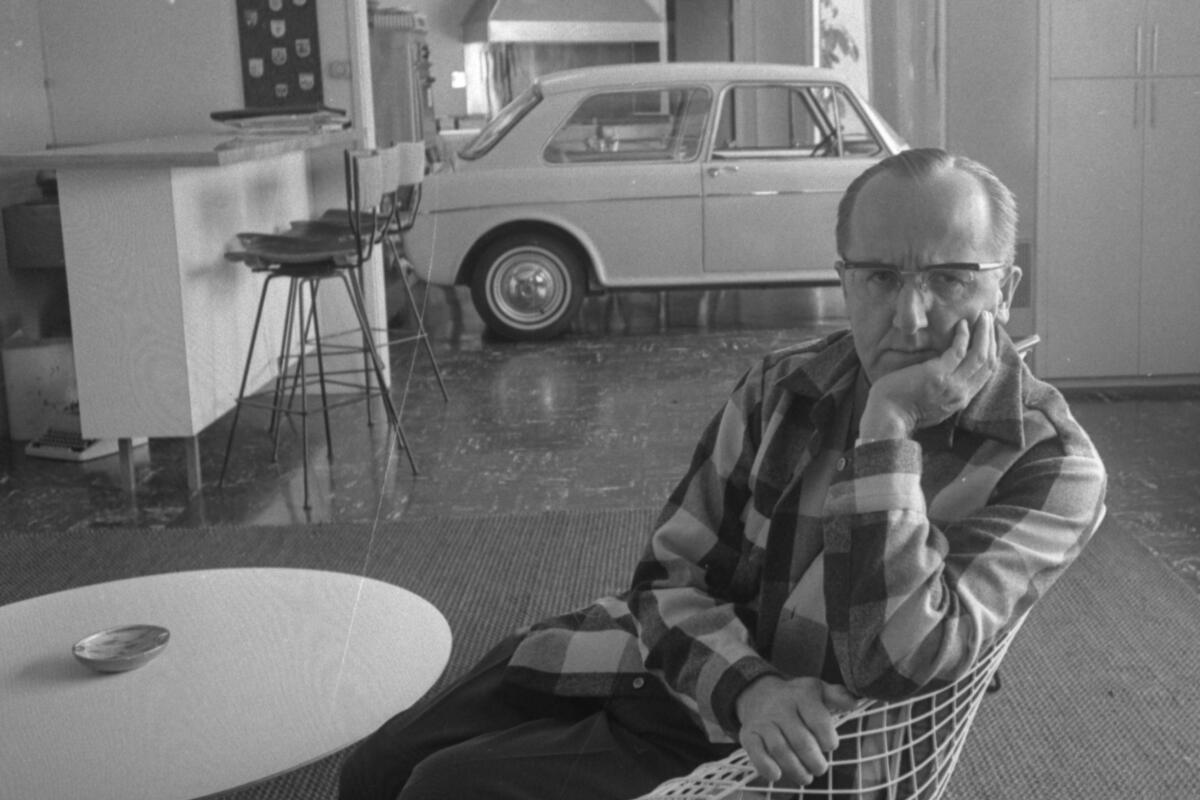 A man rests his chin on his hand and looks peeved. In the background of his home is a small car.