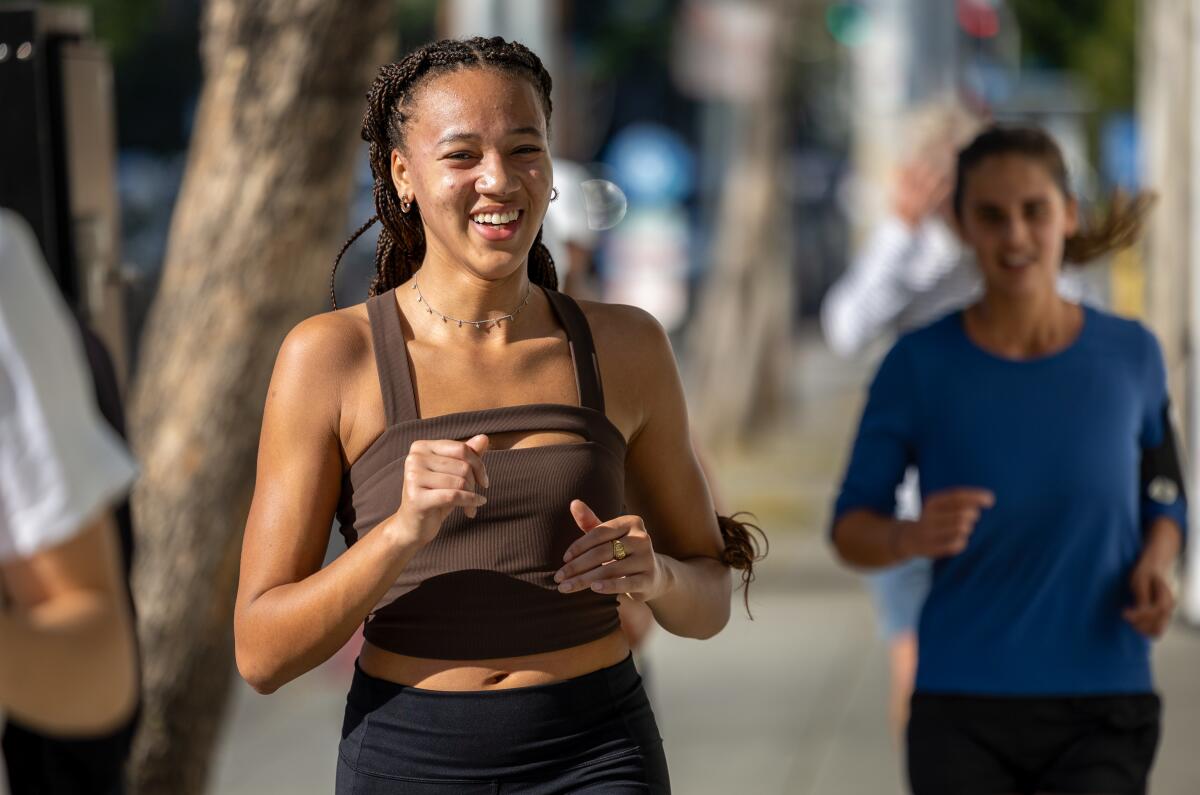 A runner smiles as she jogs on a sidewalk, flanked be fellow runners.