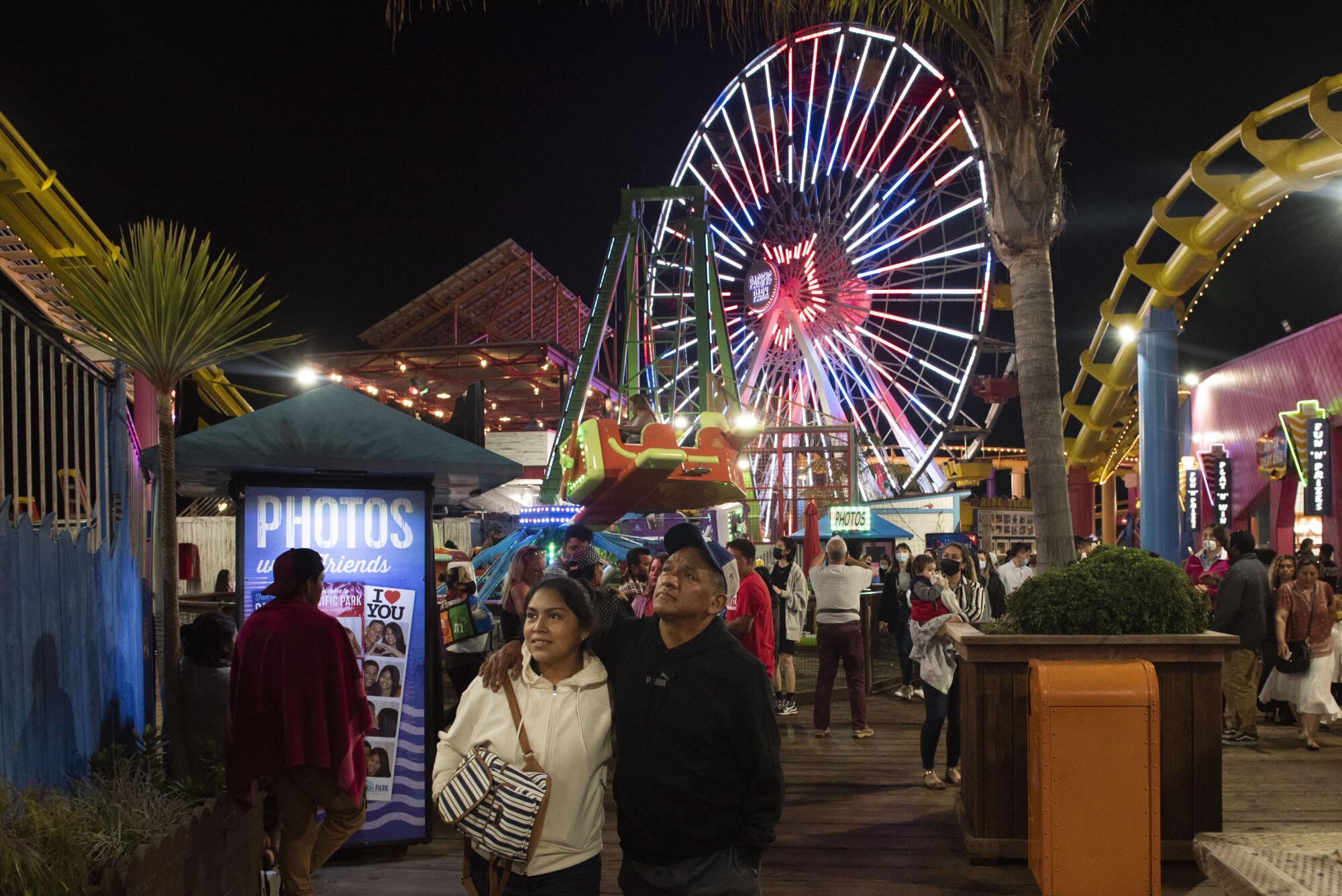 A woman and a man look up. In the background is a Ferris wheel lighted up in red, white and blue and other people.
